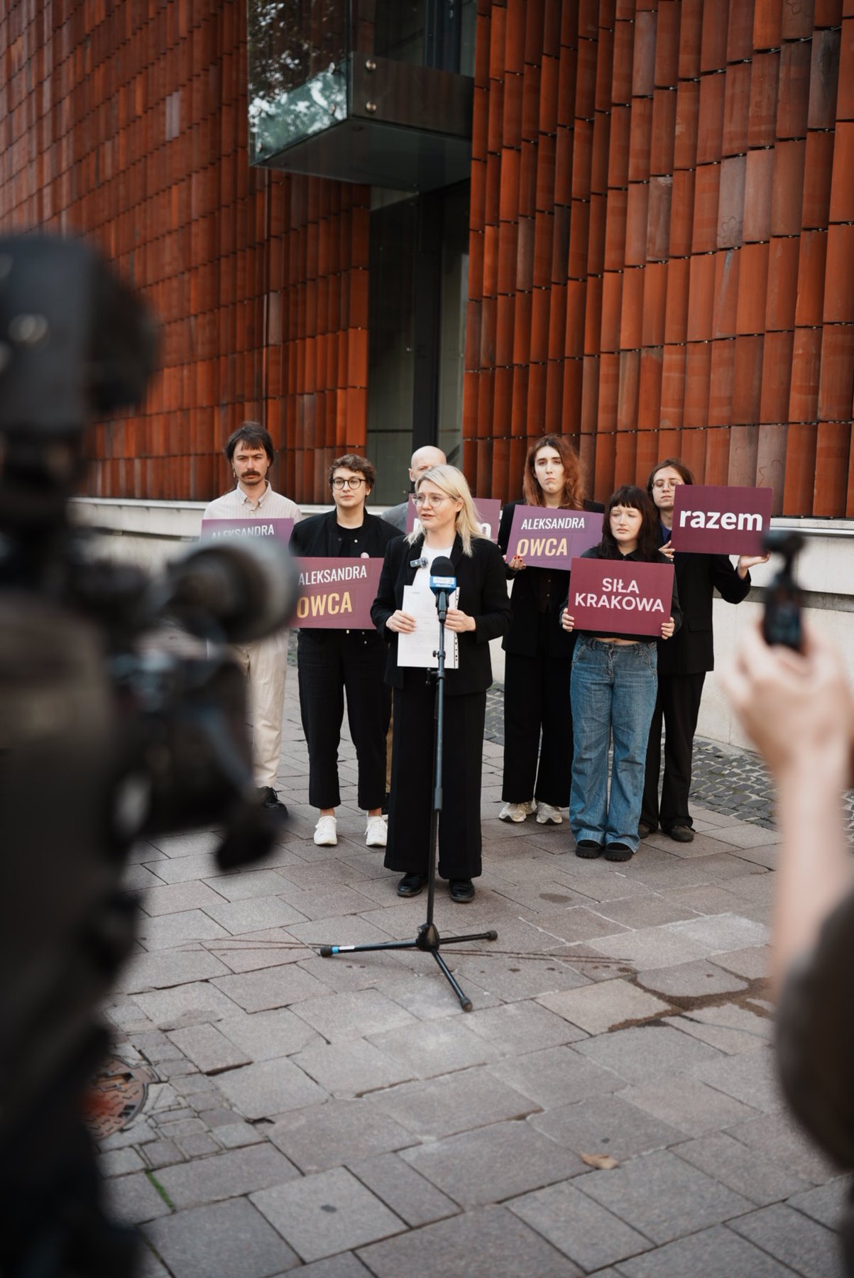 Aleksandra Owca przemawia do mikrofonu podczas konferencji prasowej Partii Razem, w tle osoby trzymające tabliczki z hasłami „Aleksandra Owca” i „Siła Krakowa”